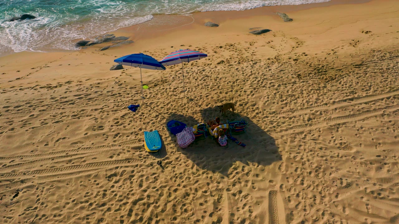 Overhead view of a beach camping setup at Shipwrecks Beach, Baja California Sur, with a blue umbrella, striped shade, gear, tire tracks, rocks, surf along the top edge, and no visible people.