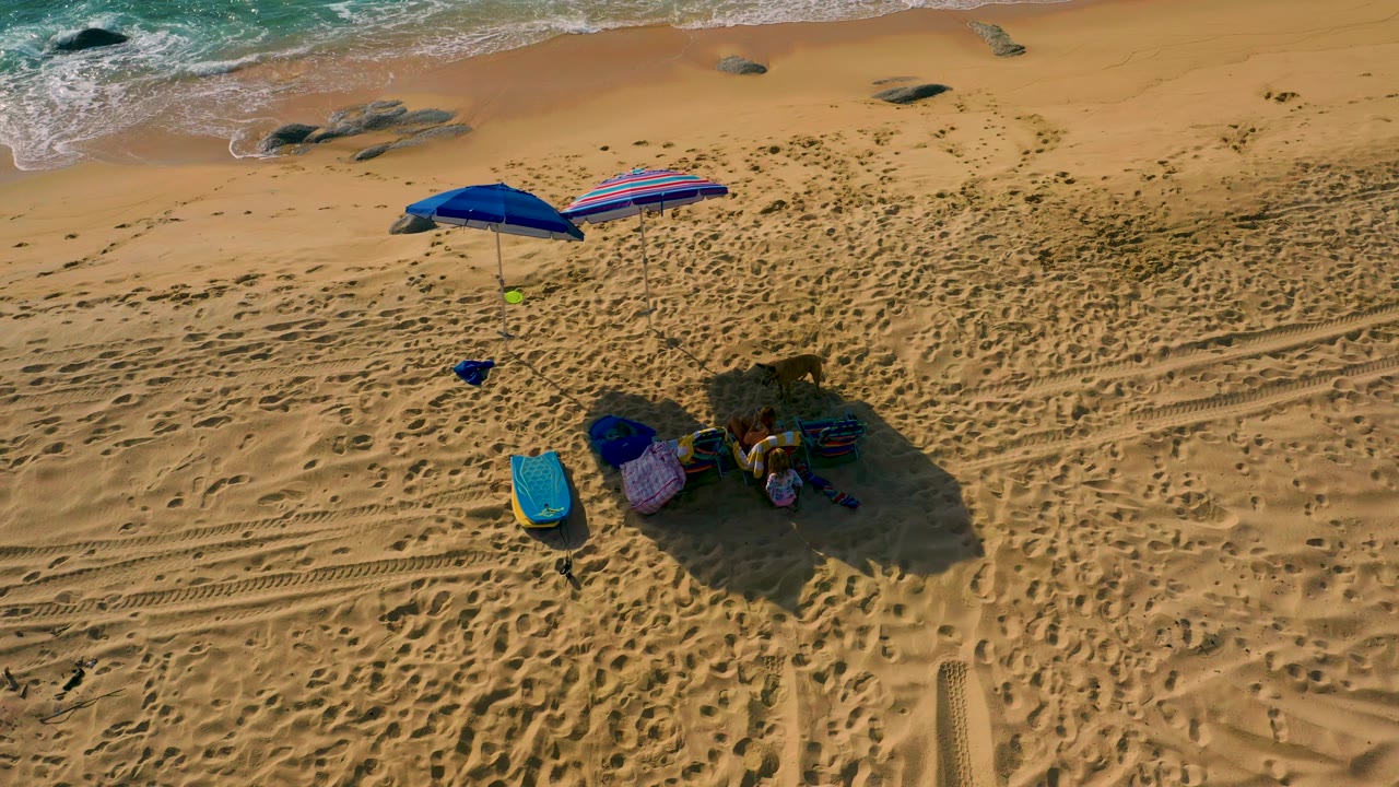 High-angle aerial view at Shipwrecks Beach, Baja California Sur, with the camp gear below center, rocks near the wet sand, foamy waves at the top, and no visible people.