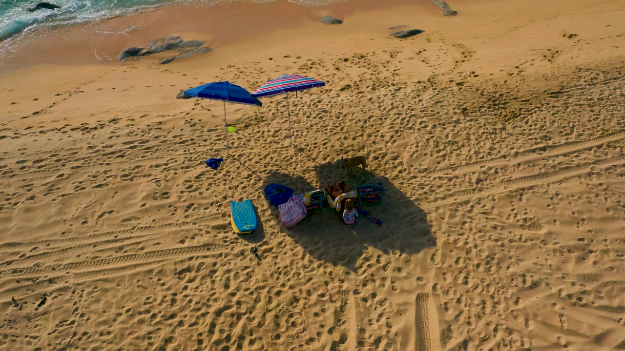 Top-down frame at Shipwrecks Beach, Baja California Sur, showing the blue umbrella, striped shade, cooler, clustered gear, long shadow, tire tracks, and no visible people.