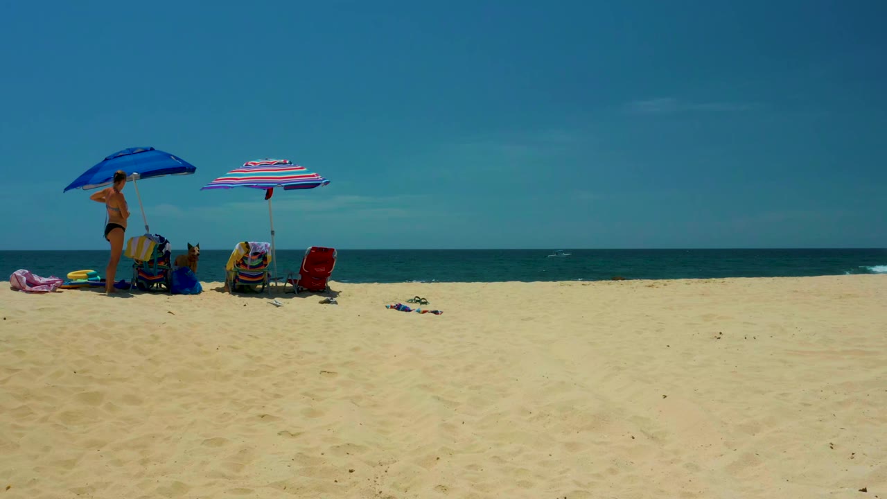 Wide beach view with two umbrellas, chairs, towels, people, ocean, and blue sky.