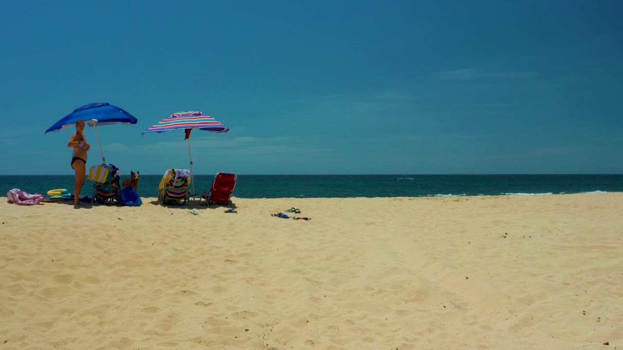 At Shipwrecks Beach in Baja California Sur, blue and pink umbrellas shade chairs and beach gear on pale sand with people standing near the shoreline and ocean behind them.