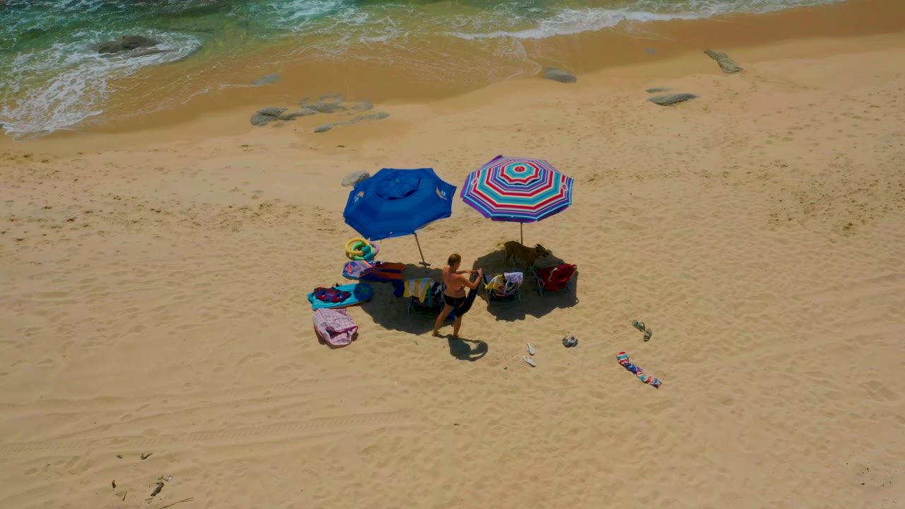 Overhead view at Shipwrecks Beach in Baja California Sur showing a blue umbrella, a striped umbrella, beach chairs, towels, and visible people on sand below the surf line.