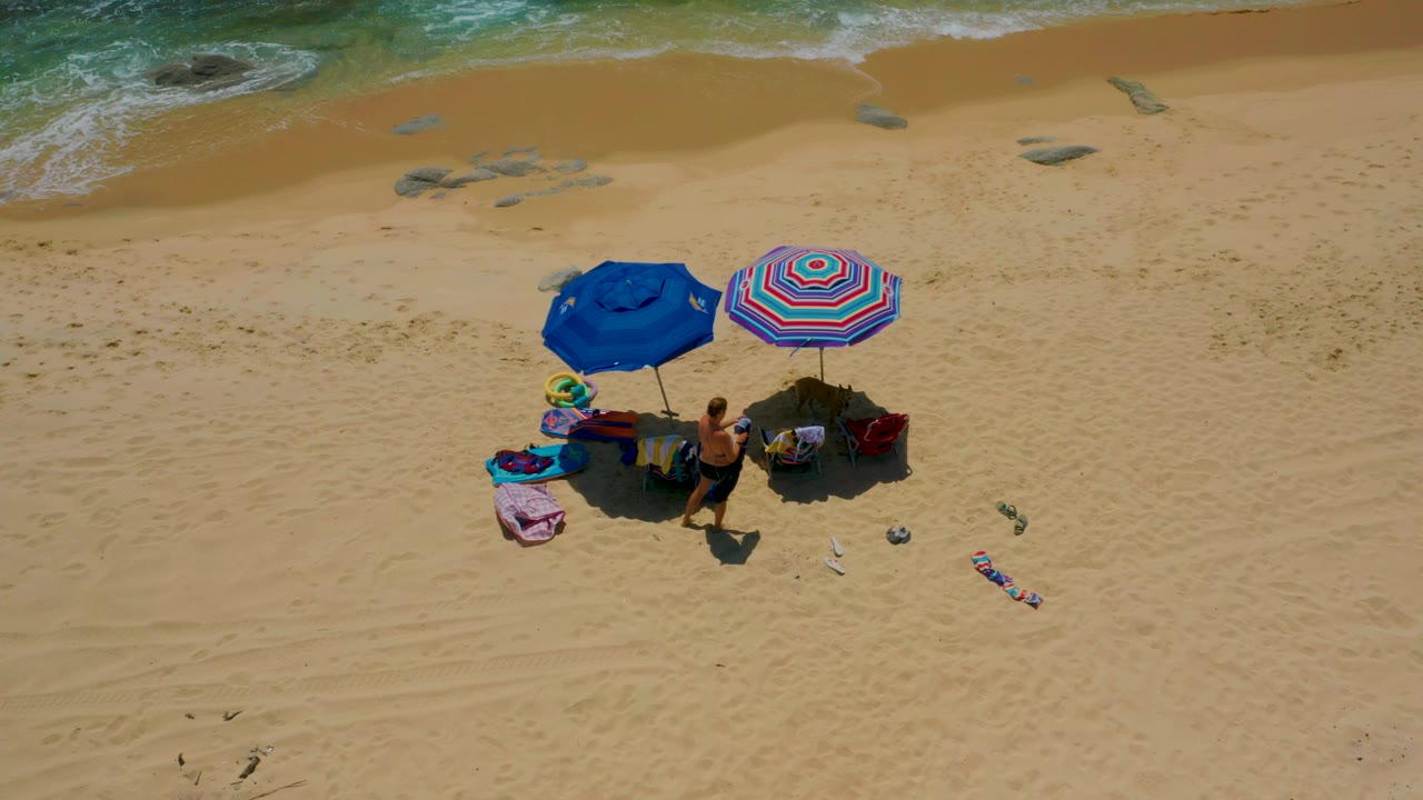 High overhead view at Shipwrecks Beach in Baja California Sur showing the umbrella pair on tan sand, several visible people near the shade, scattered gear, and surf near the top of frame.
