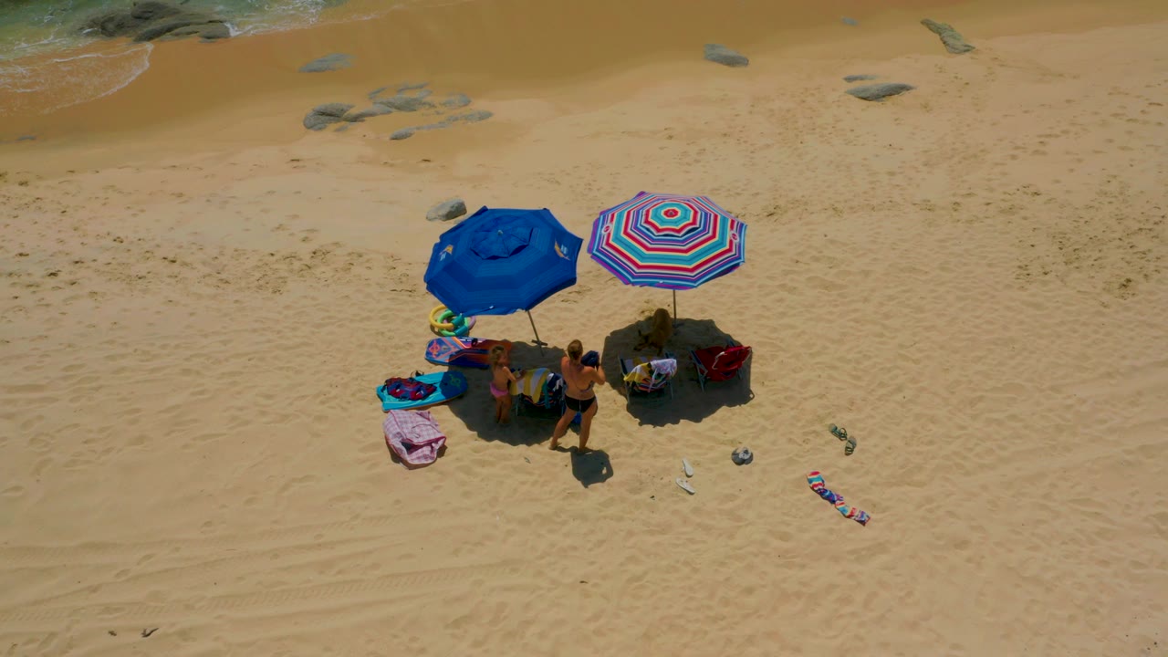 At Shipwrecks Beach in Baja California Sur, Mexico, an overhead drone frame shows blue and striped umbrellas shading visible beachgoers, chairs, towels, and scattered sandals on tan sand near a thin line of surf.