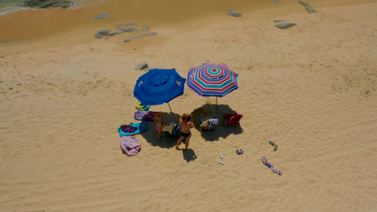 At 0:02, the aerial composition at Shipwrecks Beach shows visible beachgoers clustered beneath two umbrellas, with a towel and beach items extending outward across the sand and shallow water along the top edge.