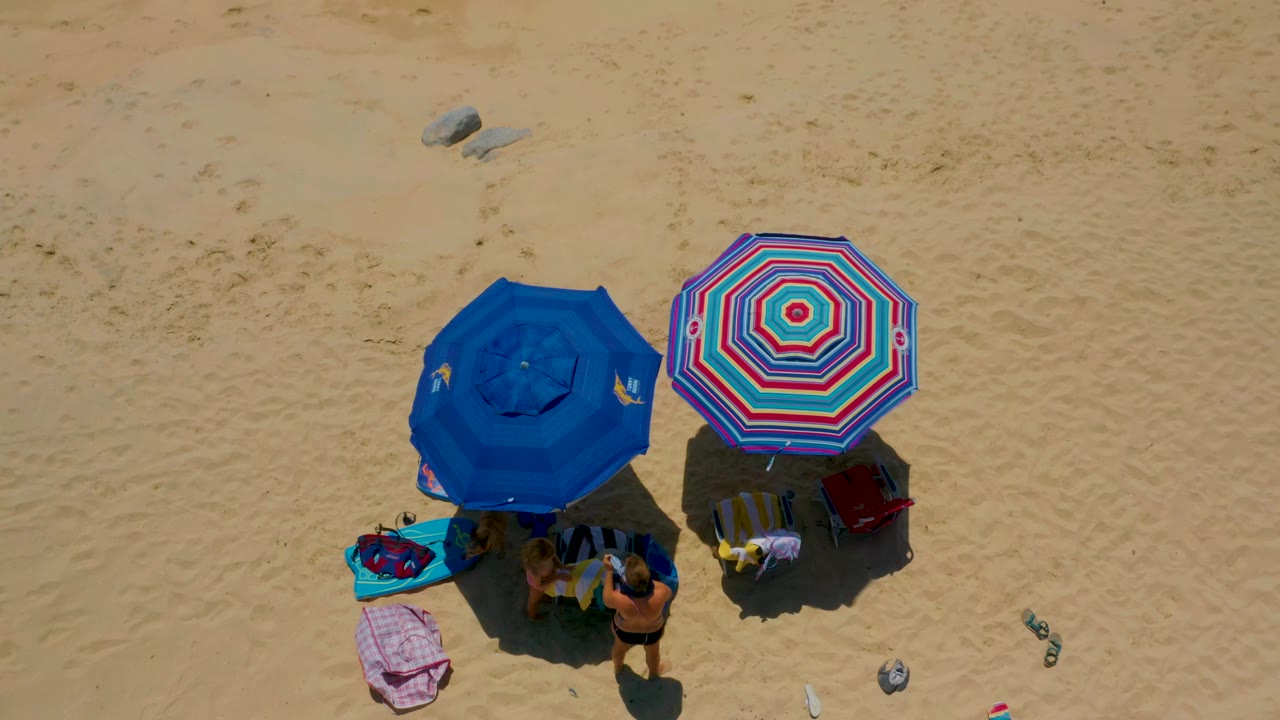 At 0:01, the Shipwrecks Beach overhead view keeps the blue and striped umbrellas centered with towels, bags, sandals, bottles, rocks, and people partly hidden below the shade.