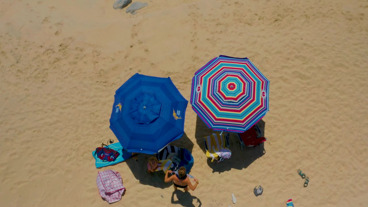 At 0:03, the Shipwrecks Beach aerial view places the umbrellas larger in frame, with a person near the lower edge, towels, bags, sandals, bottles, and small rocks on sunlit sand.