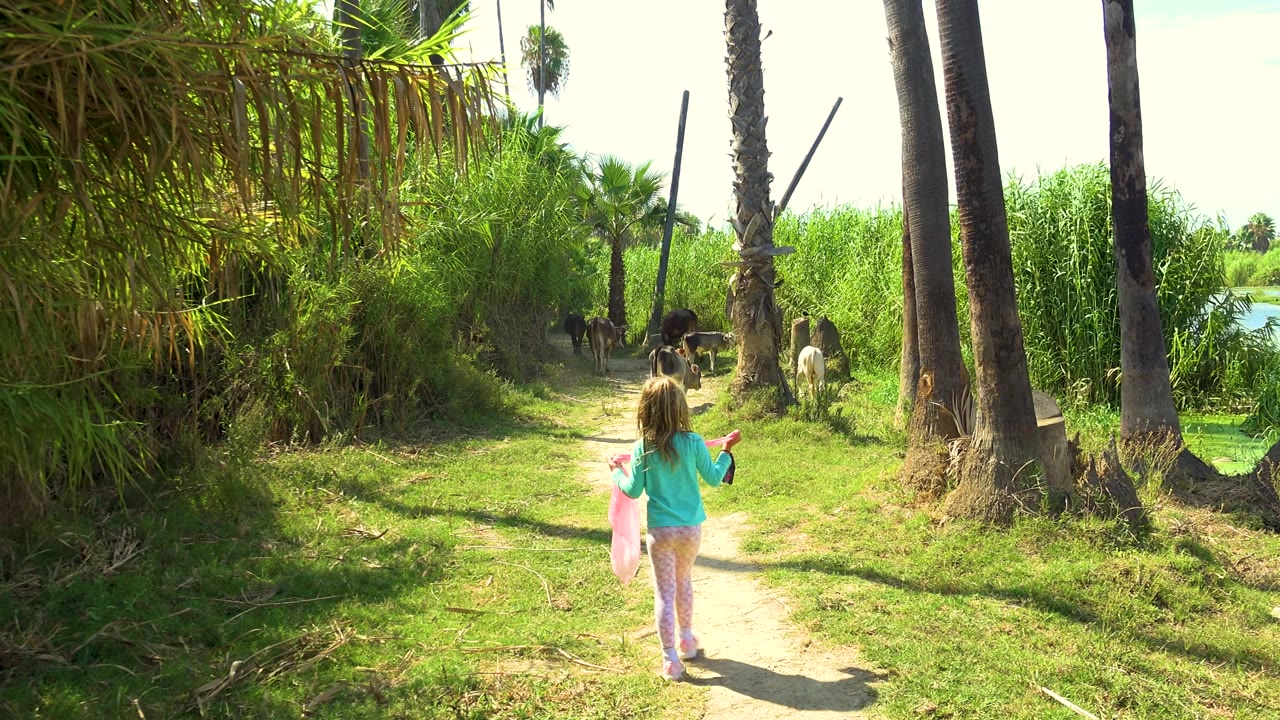 The girl moves slightly to the right side of the path, arms lifted with a pink item visible, while cattle remain ahead among palms and thick greenery.