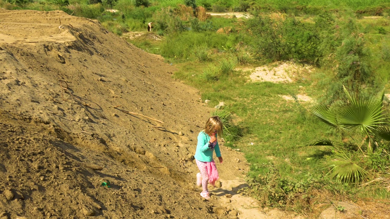 A young girl in a turquoise shirt and pink pants walks near the base of a steep sandy embankment with green brush and a small palm nearby.