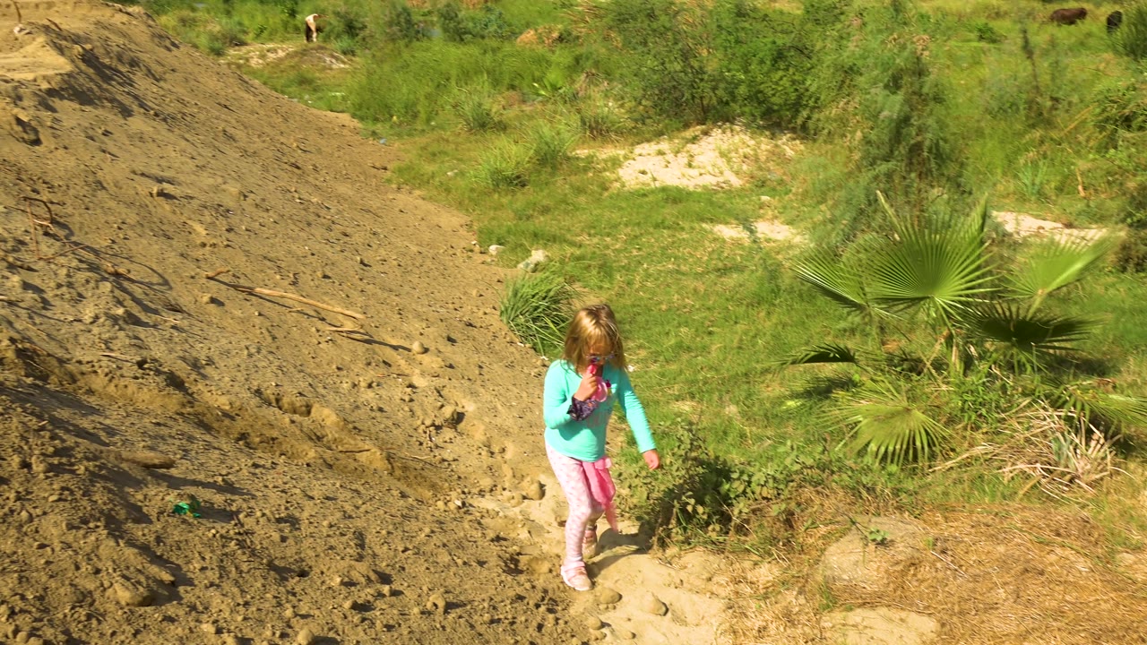 The child moves farther right in frame on the dirt path, passing low vegetation and a fan-shaped palm under strong sunlight.