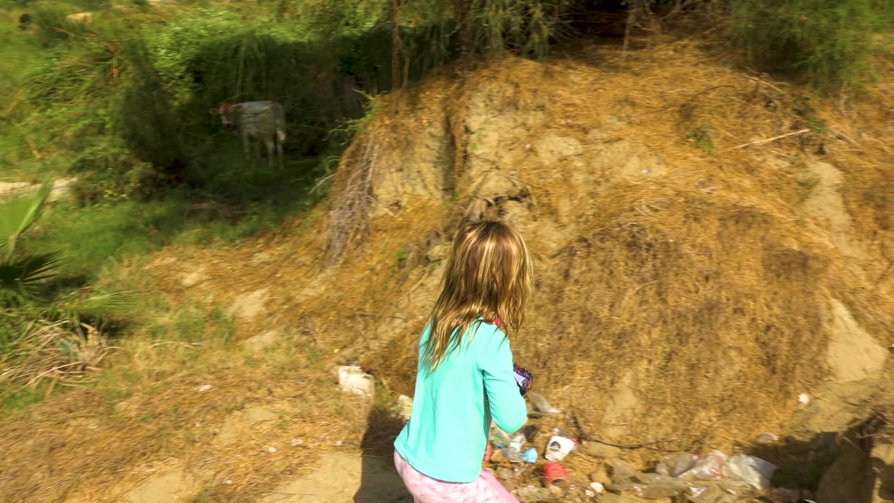 A blonde girl in a teal shirt and pink pants stands near scattered debris at the base of a dry eroded bank, with green brush above the path.