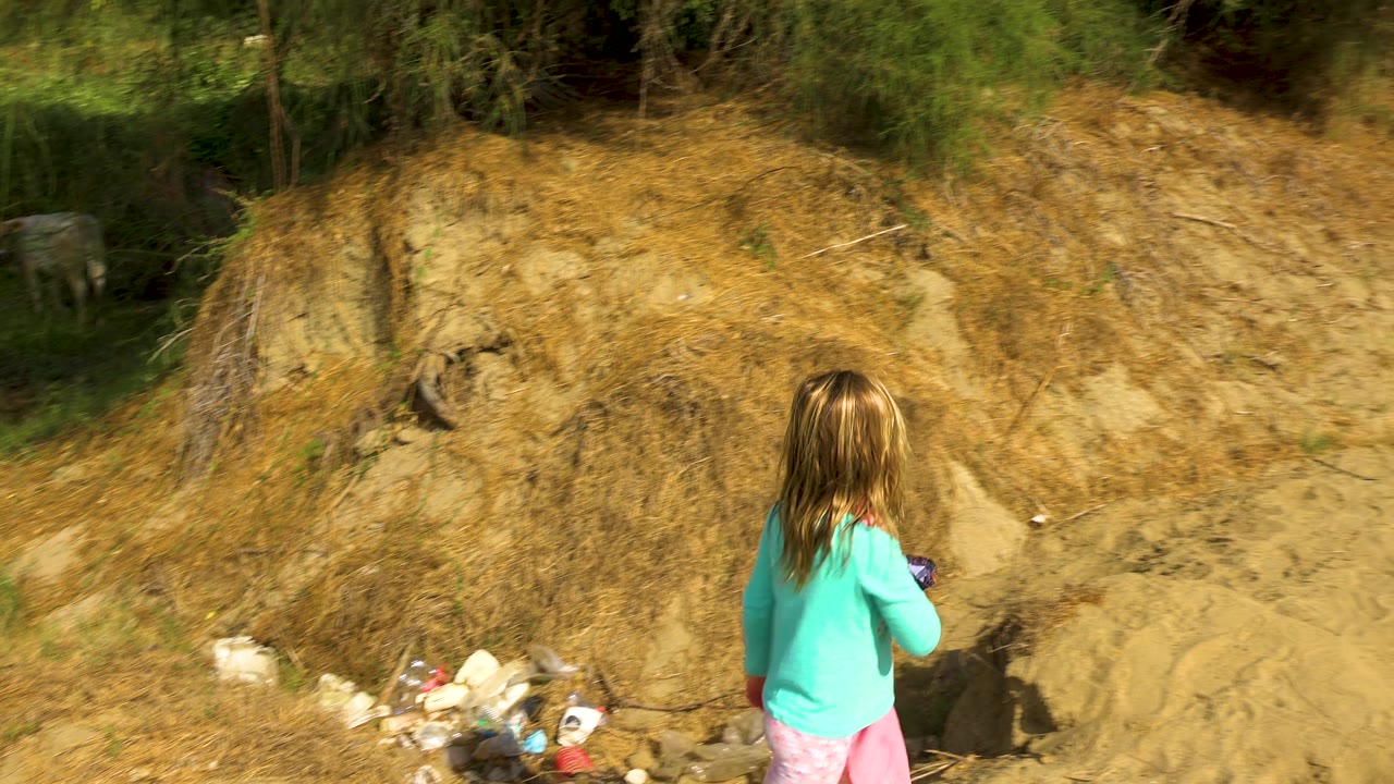 The child continues away from camera on the sandy trail, moving past debris toward a path bordered by dry banks and green brush under bright sky.