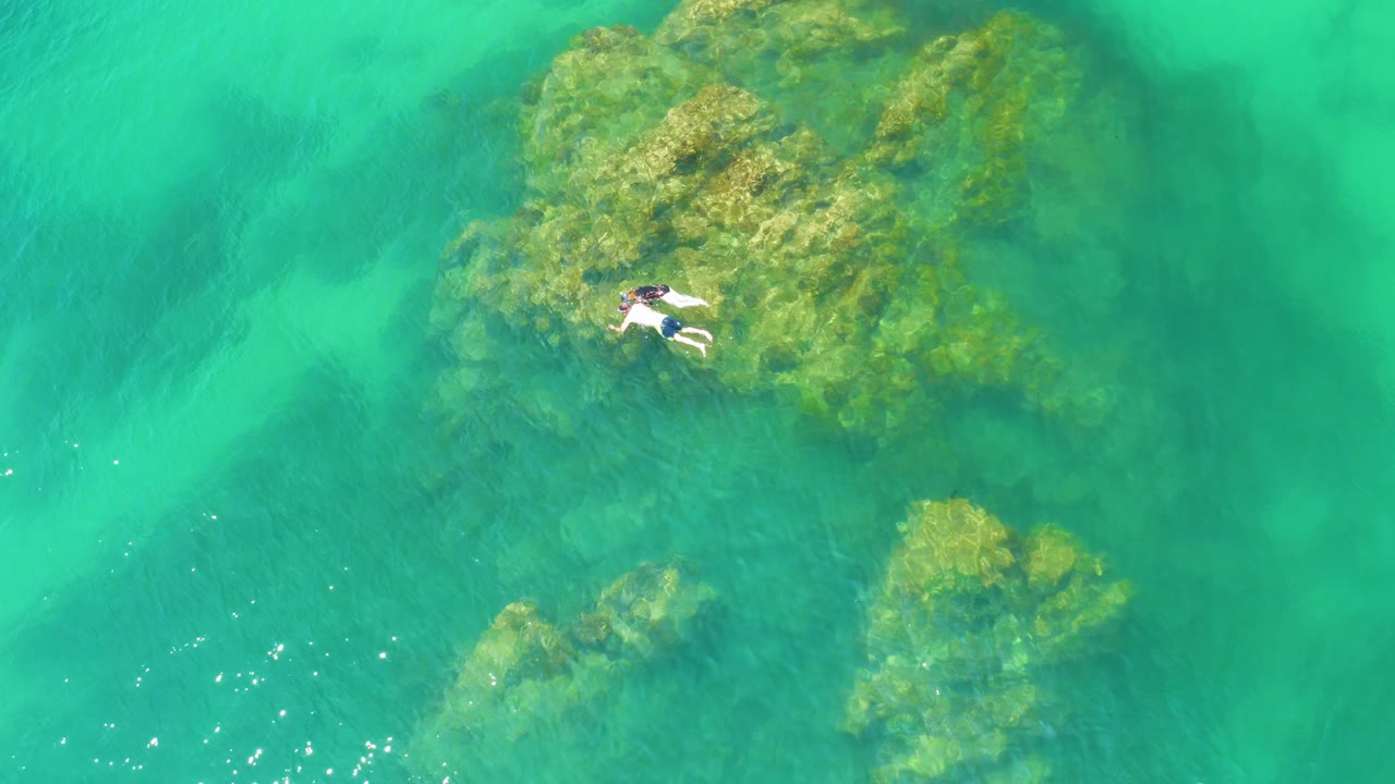 At 0:02, the snorkeler remains small in the frame above the upper reef, with deeper green water texture filling the surrounding view.