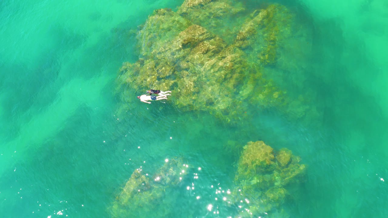 Overhead view of two snorkelers beside submerged reef rocks in turquoise water.