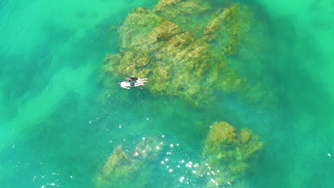 At 0:01, the Palmilla Beach aerial view keeps two small snorkelers near the upper reef patch while smaller algae-covered rocks and scattered light reflections sit lower in the frame.