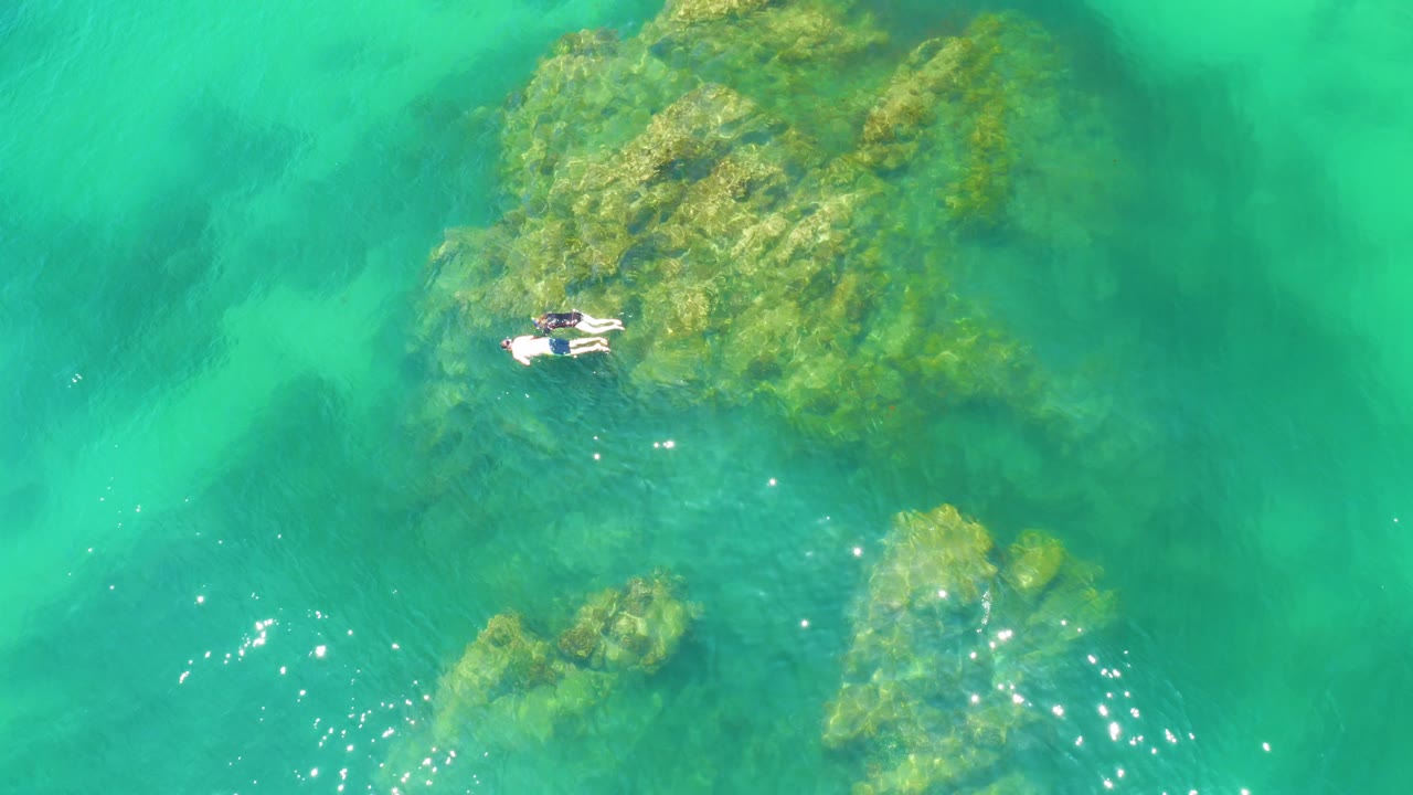 At 0:02, two distant snorkelers are positioned along the left edge of the large reef area at Palmilla Beach, with clear green-blue water and a smaller rock cluster below.