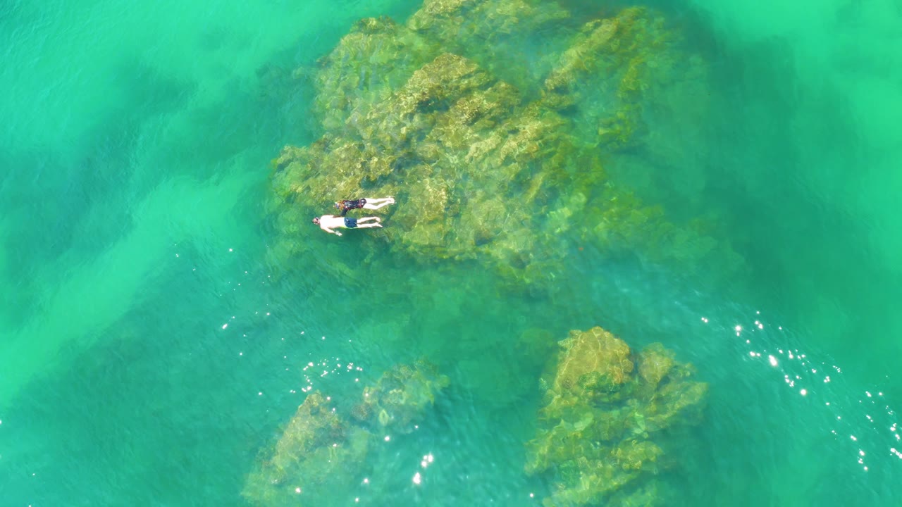 At 0:03, the Palmilla Beach drone view shows two distant snorkelers close to the left side of a broad algae-covered rock, with open turquoise water filling the right side.