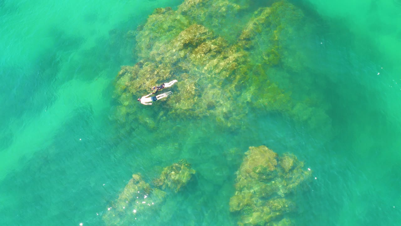Overhead 4K frame from Palmilla Beach showing a lone swimmer floating beside a large algae-covered rock formation in bright turquoise water.