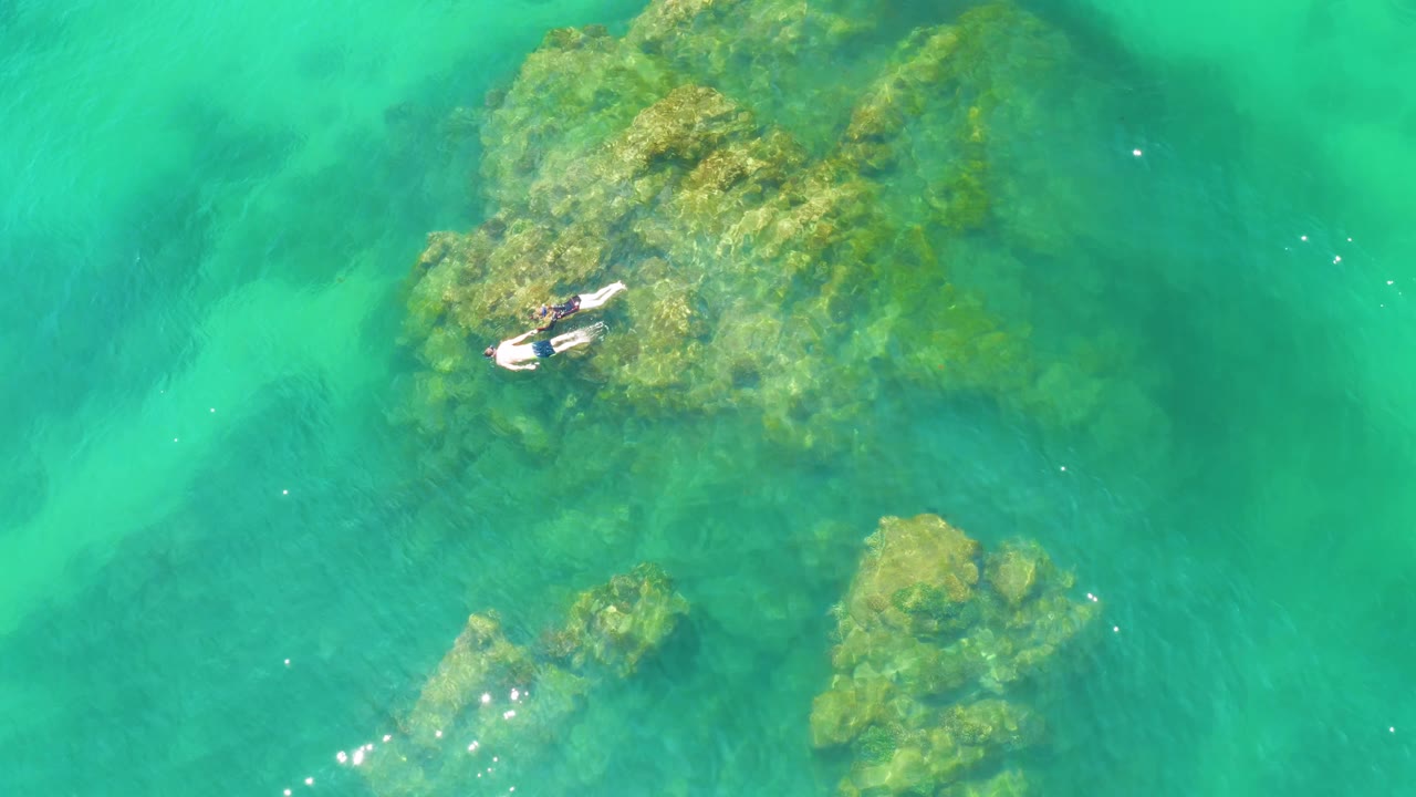 Swimmer floats near a rocky patch under clear green-blue water.