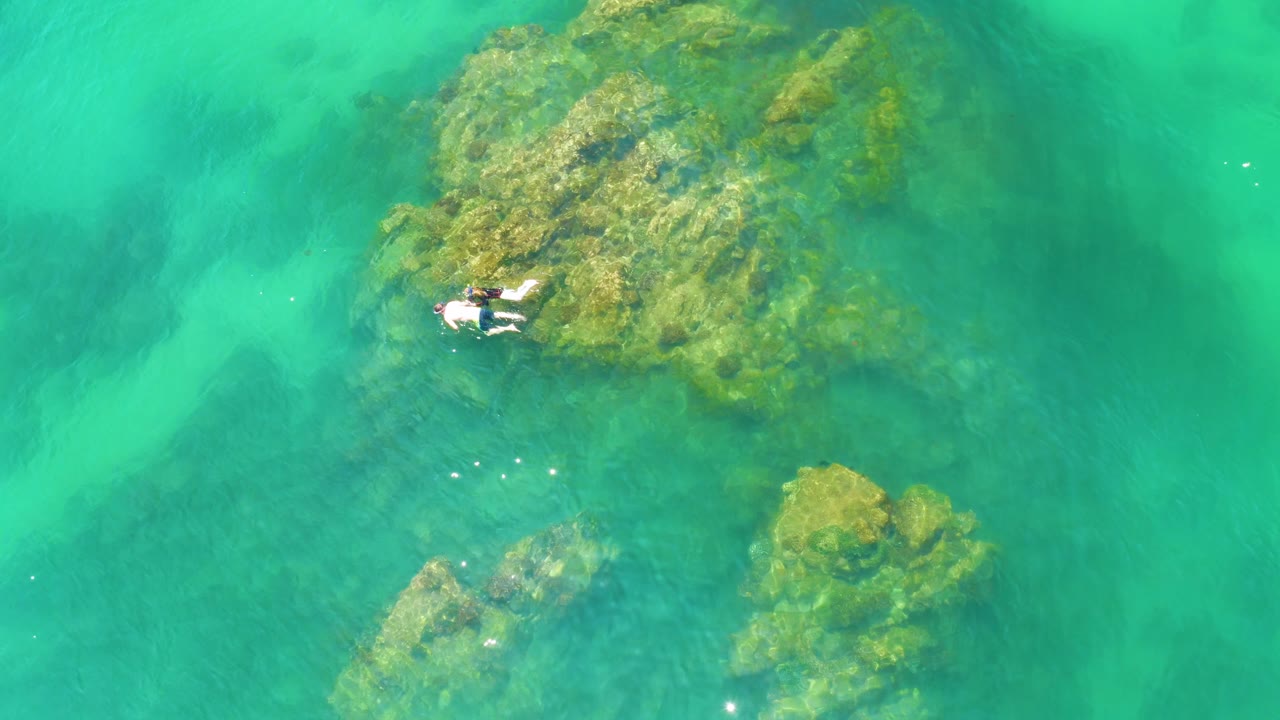 The overhead frame shows the swimmer slightly farther along the rock formation at Palmilla Beach, with darker green patches under the water nearby.