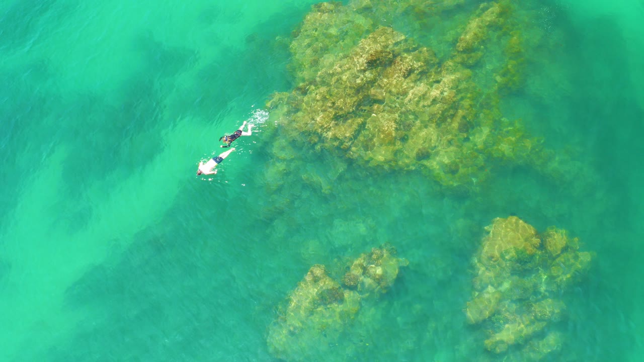 At Palmilla Beach, Baja California Sur, two distant snorkelers float near the upper left of clear turquoise water beside yellow-green submerged reef.