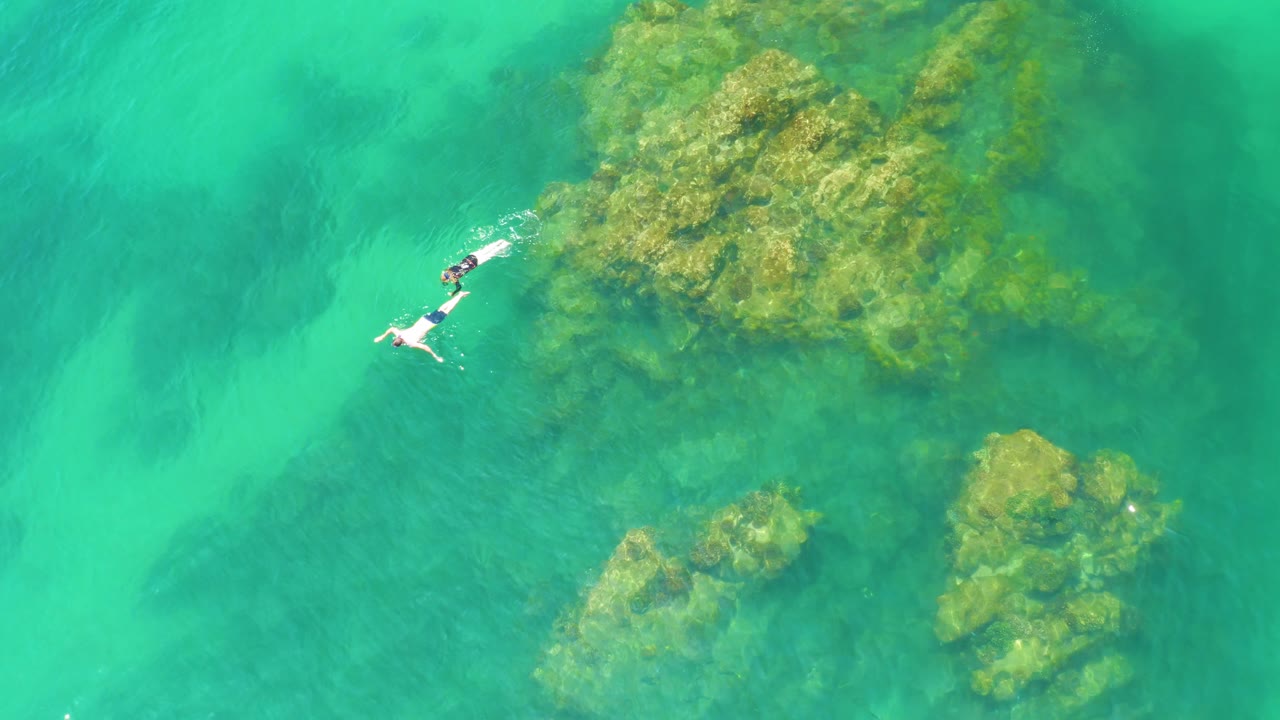 At 0:02, the snorkelers remain near the left side of frame while a broad reef mass and smaller rock clusters show through the green-blue water.