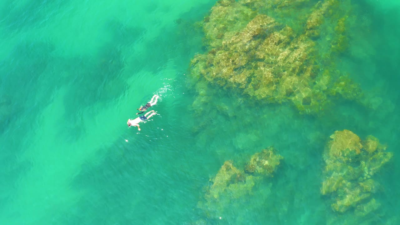 At Palmilla Beach in Baja California Sur, the two snorkelers appear near the upper left while translucent green-blue water reveals a large rocky area on the right and smaller rocks below.
