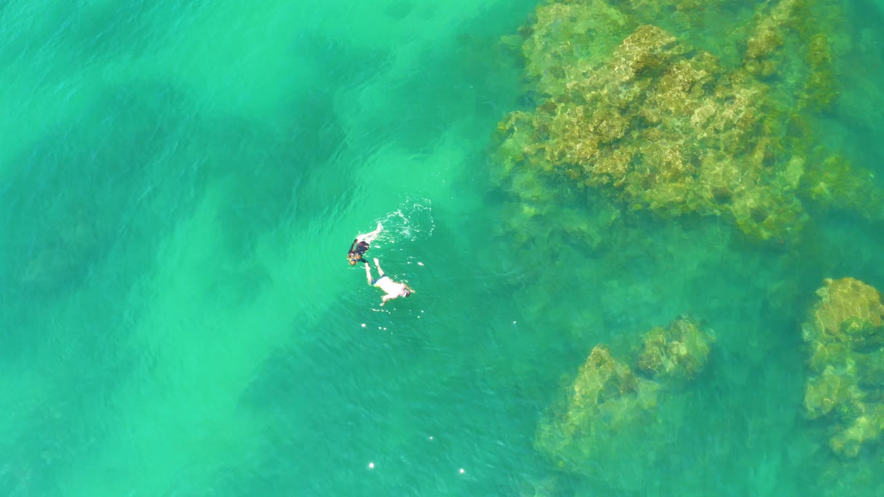 At Palmilla Beach, two finned snorkelers float in bright green-blue water while a textured submerged rock mass fills the upper right of the frame.