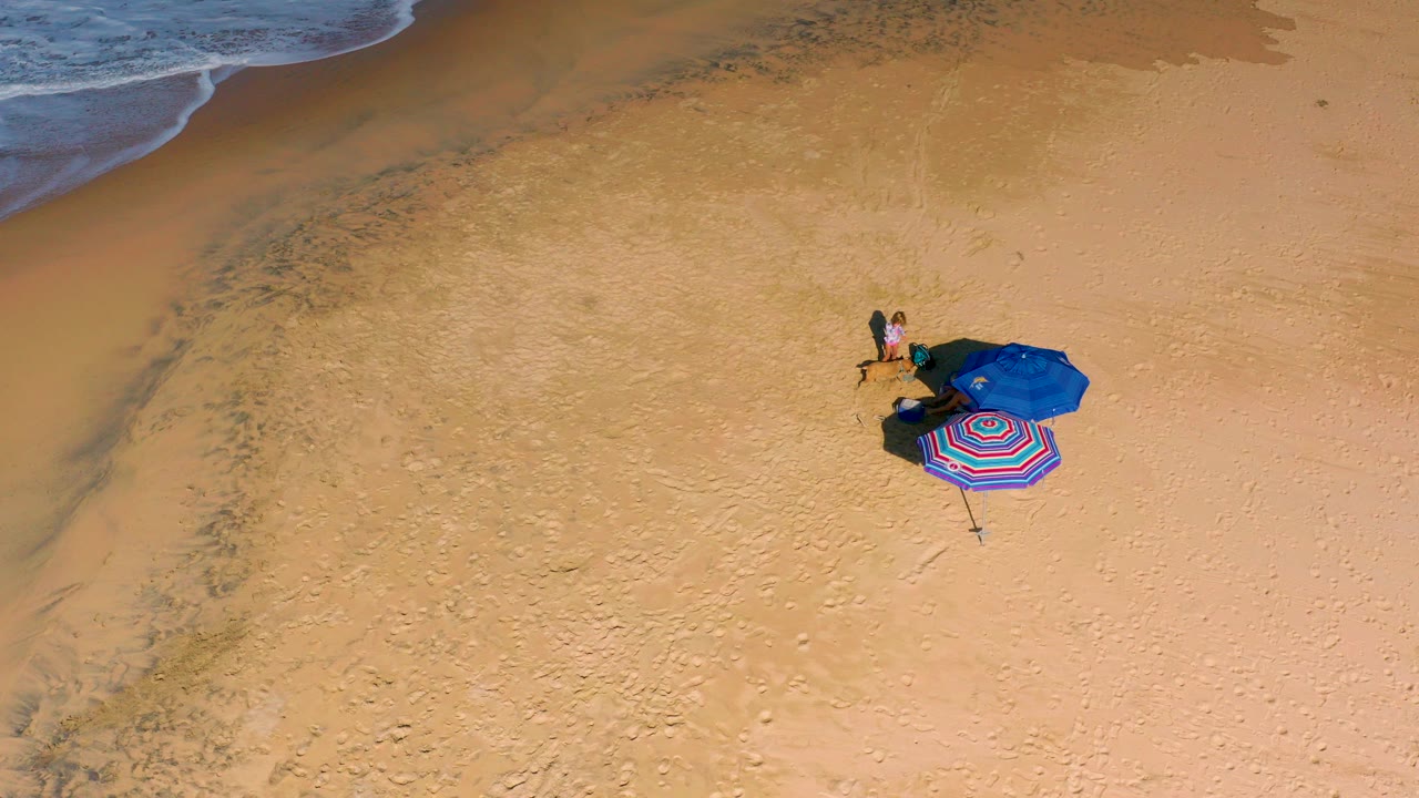 Top-down aerial view of a sandy beach with the shoreline at upper left, a blue umbrella, a striped umbrella or towel, and tiny people near the setup.