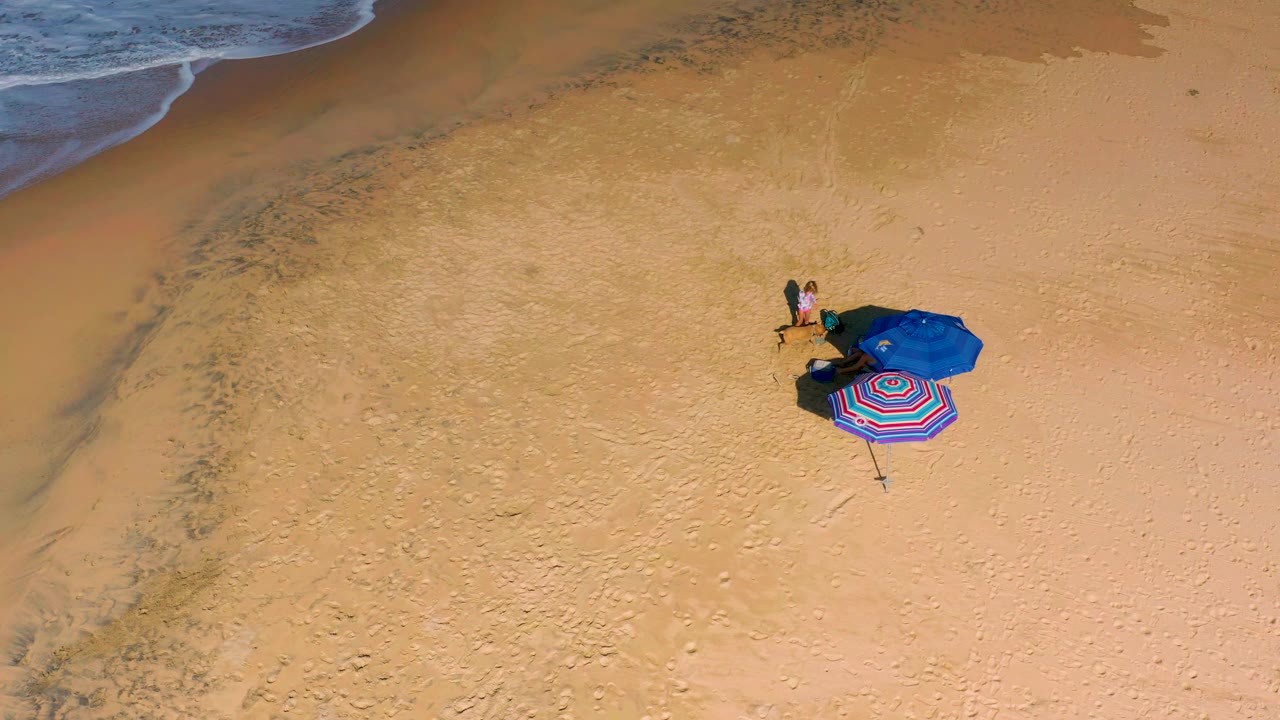 Overhead drone frame showing a small beach setup on open sand, with a blue umbrella, striped beach item, dark shadows, and tiny people beside it.