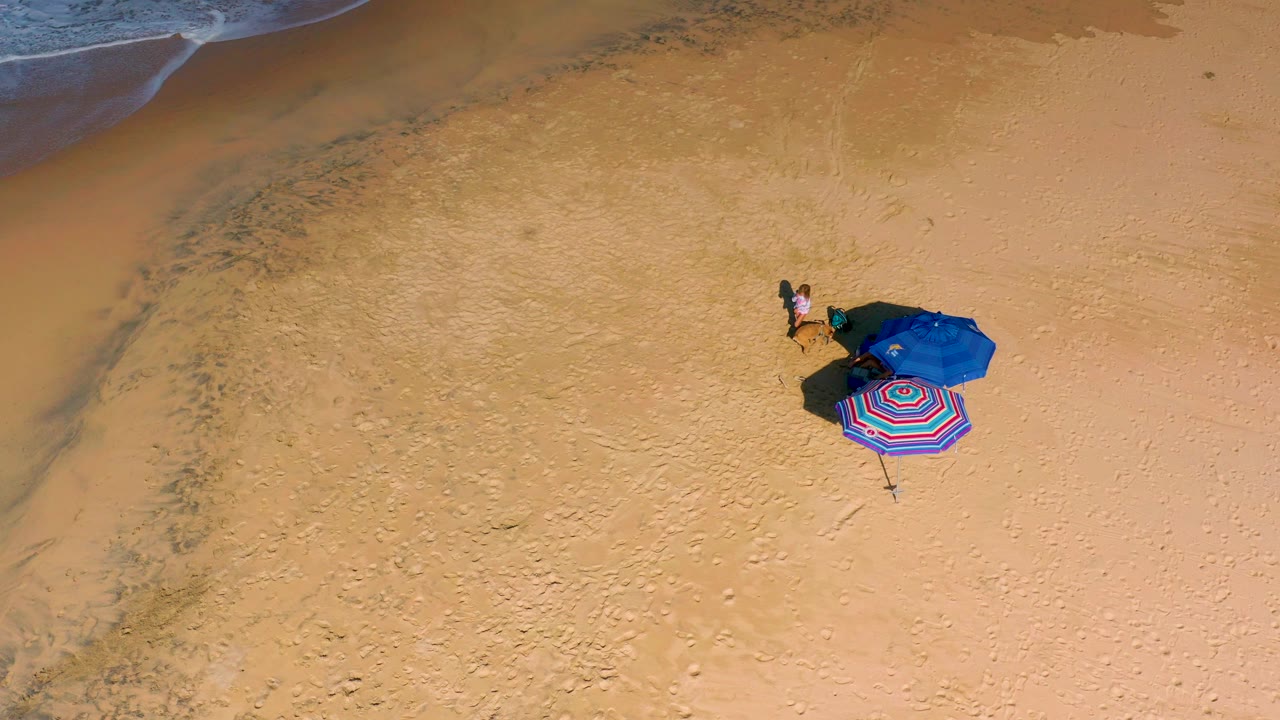 Top-down beach view with the ocean edge partly visible at upper left, a clustered umbrella and towel setup to the right, and tiny people standing nearby.