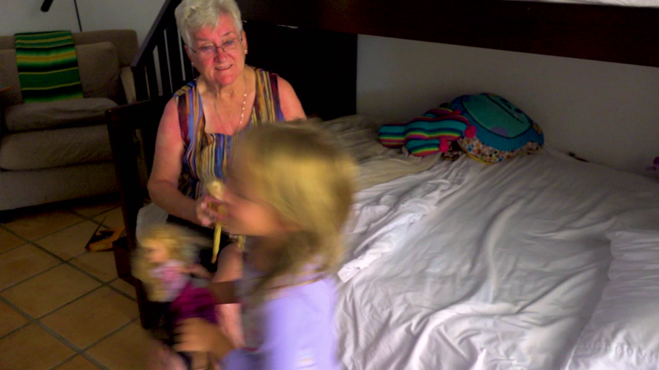 An elderly woman with short white hair sits on the edge of a bed while a young child in purple moves across the foreground holding a doll in a bedroom.