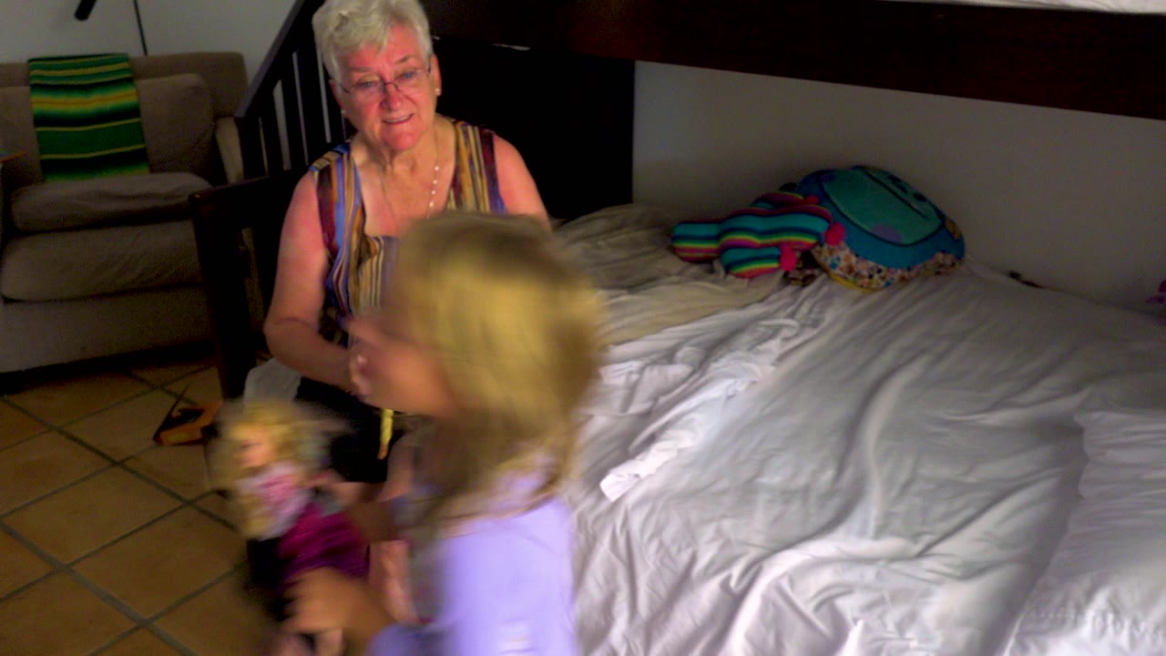 Child in purple pajamas crosses in front of grandmother sitting on the bed.