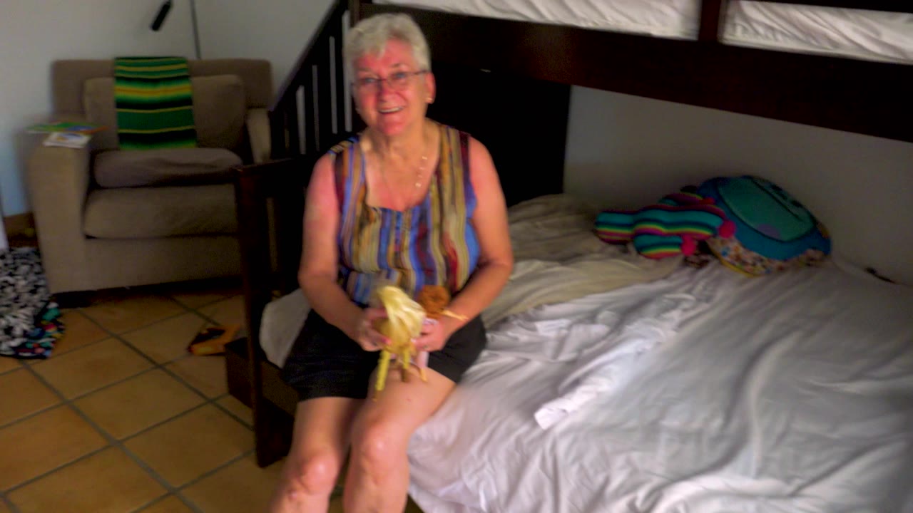 The grandmother sits centered on the bed smiling and holding dolls, with white bedding, an armchair, tiled floor, and a bunk bed visible around her.