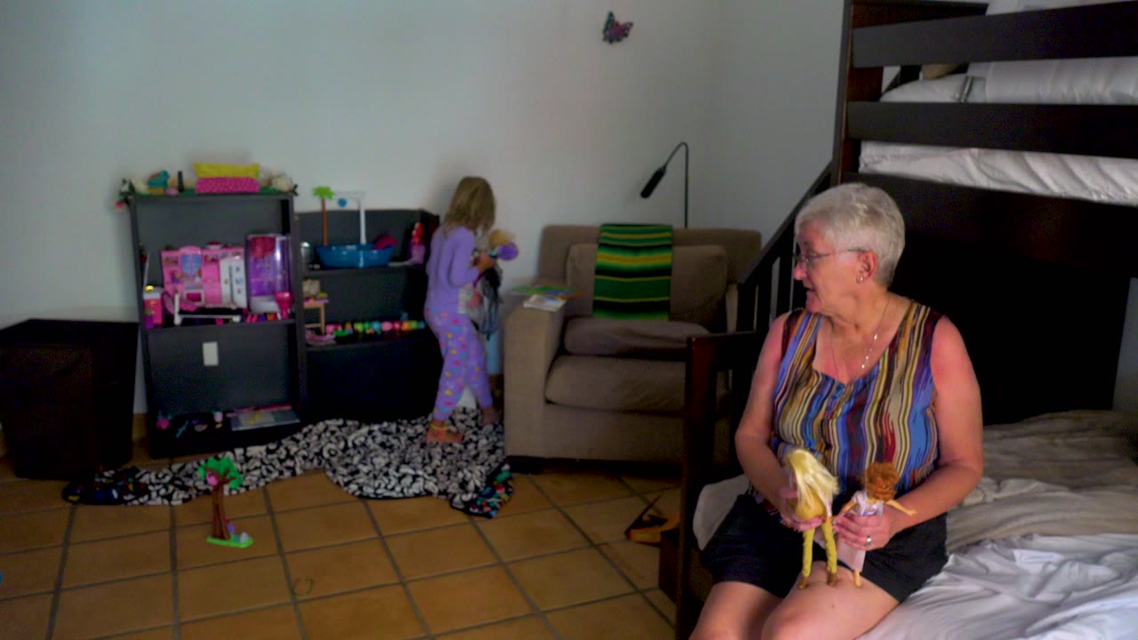 An elderly woman with short white hair sits on the edge of a bed holding a peeled banana while a child in purple pajamas stands near toy shelves in a bedroom.