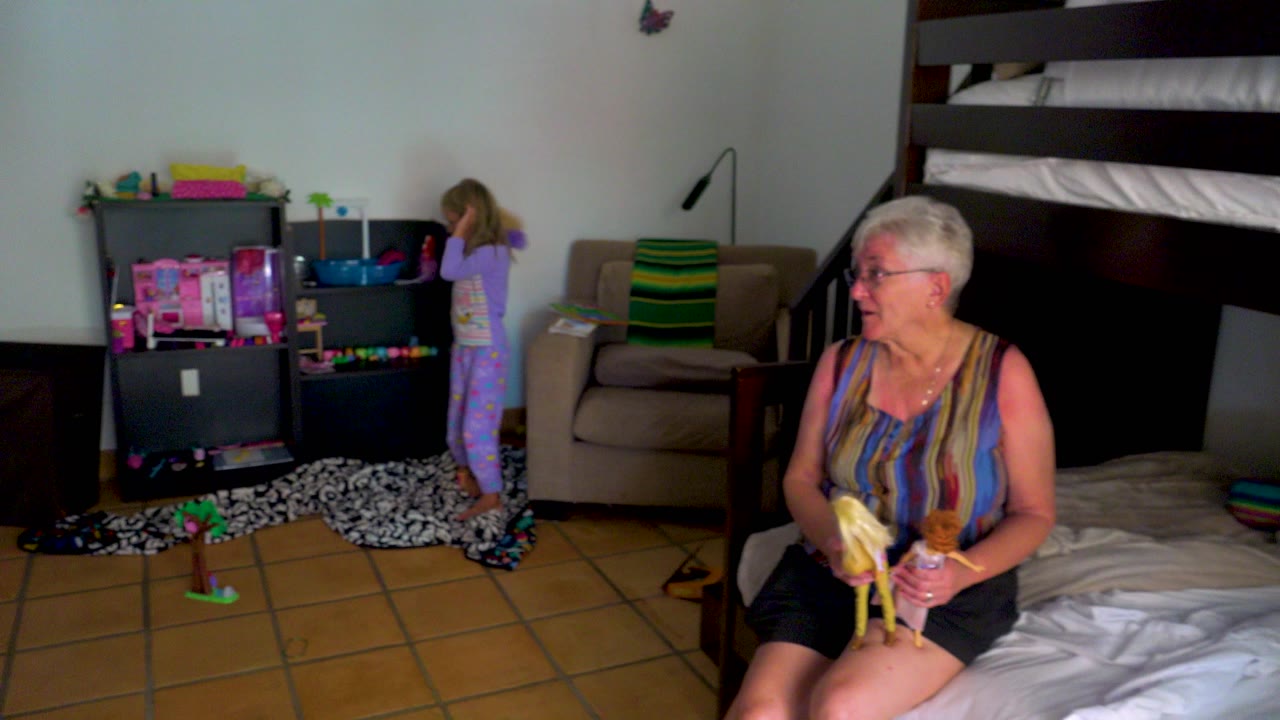 An elderly woman with short white hair sits on the edge of a bed holding a banana and doll while a child in purple pajamas faces a black toy shelf in a bedroom.