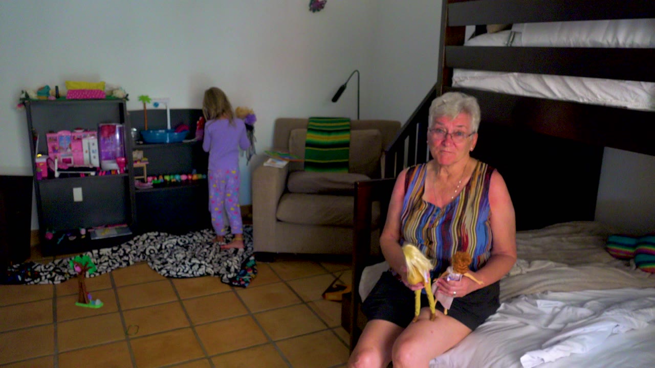At the start of the clip, an elderly woman sits on a bed holding dolls while a young girl in purple pajamas faces a toy shelf in a bedroom.