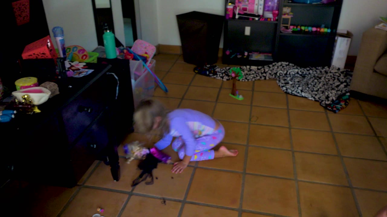 Toddler kneels near a doll beside a black cabinet in a living room.