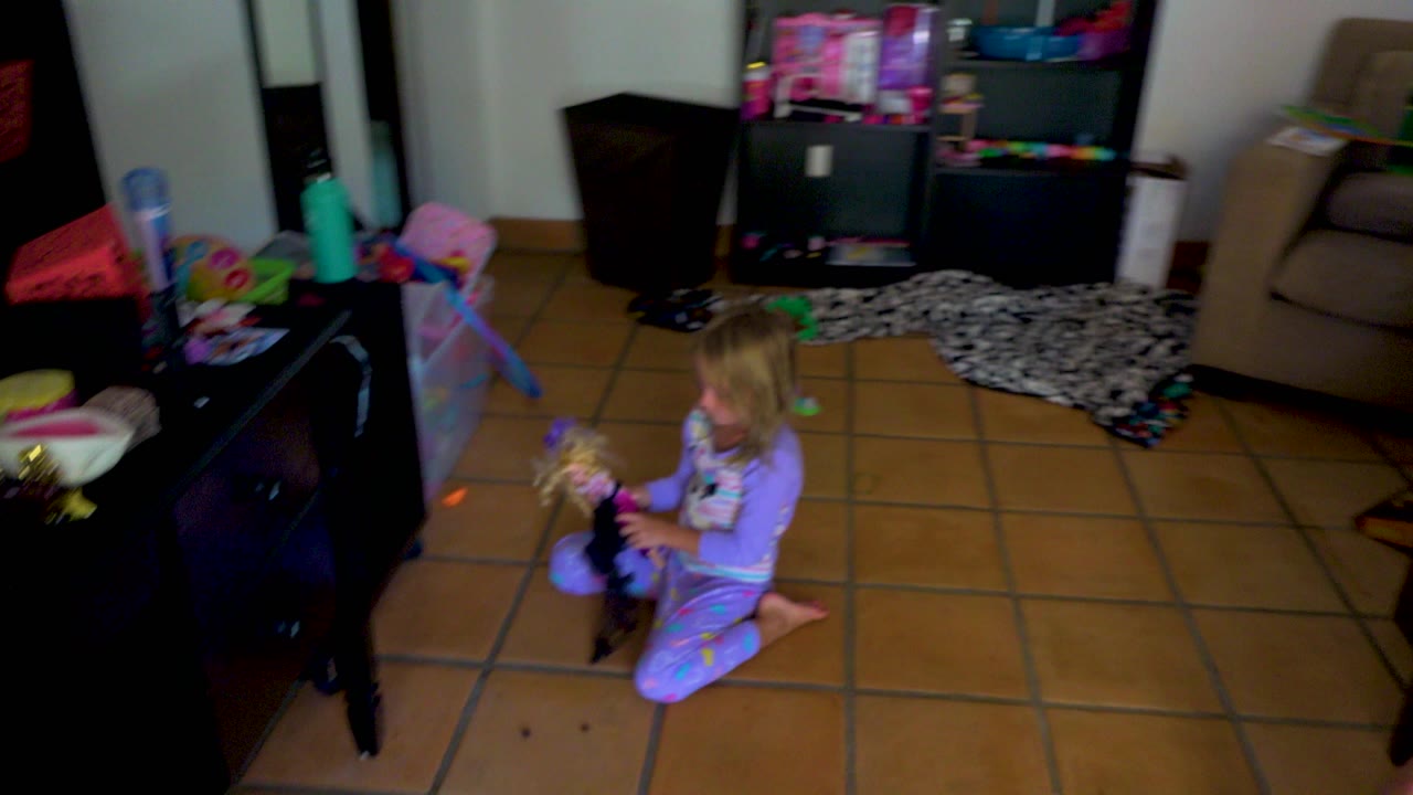 A blonde toddler sits upright with the doll in her lap on the tiled living room floor, with toy shelves, a patterned rug, and a beige armchair behind her.