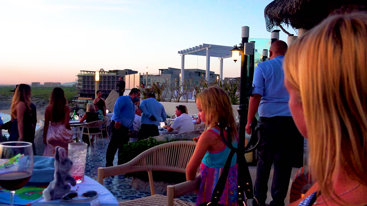 Child beside a patio table as adults dine near the beach.