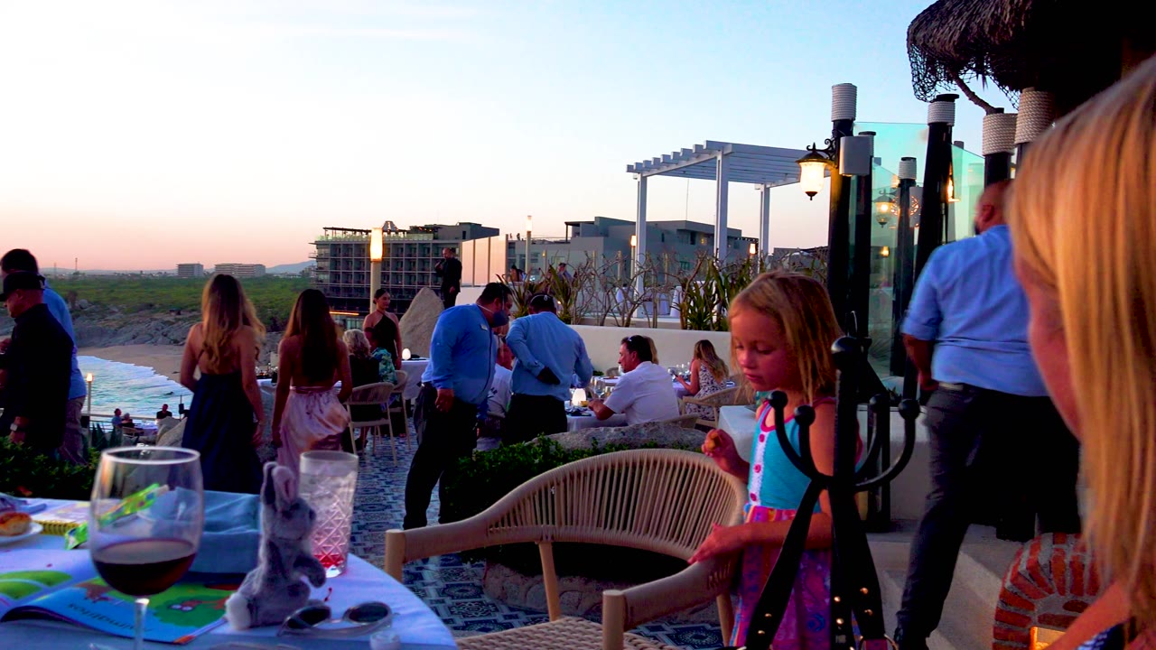 A young girl eats at the right side of the frame as patio diners gather near the terrace edge, with a wine glass and table items in the foreground and ocean visible beyond.