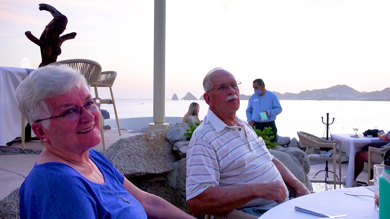 An elderly woman in a blue top smiles in the foreground while an elderly man in a striped shirt sits beside a white restaurant table with the ocean and distant islands behind them.