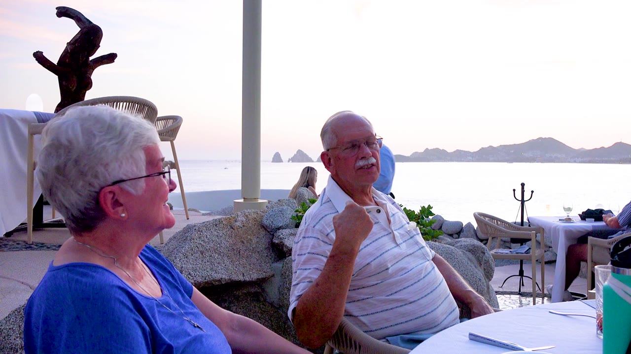 The elderly woman is shown in profile facing the elderly man, who raises one hand while seated at the white table, with outdoor dining furniture, rocks, bright ocean, and low islands behind them.