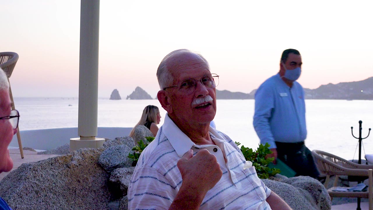 A senior man with glasses and a white mustache laughs near an outdoor restaurant table, with ocean water and rocky islands visible at dusk behind him.