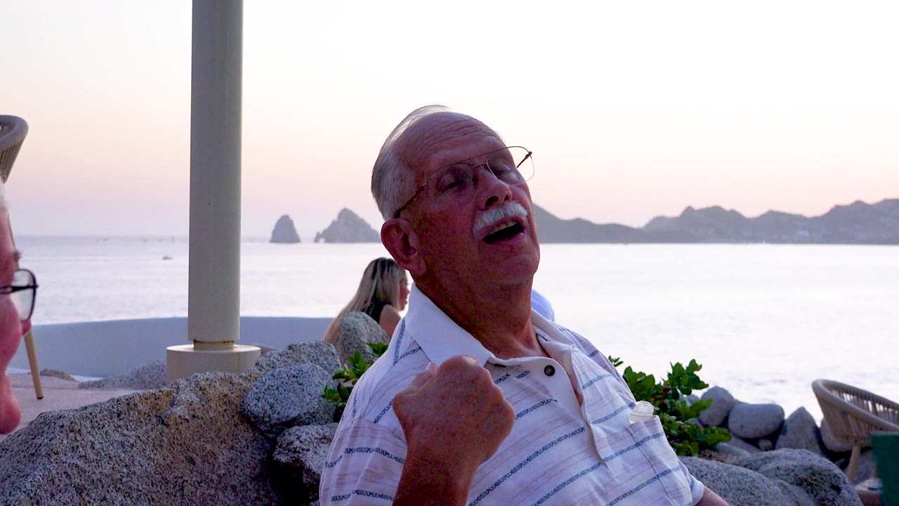 The man leans back laughing, his hand near his chest, with the calm ocean and silhouetted rocky islands filling the background.