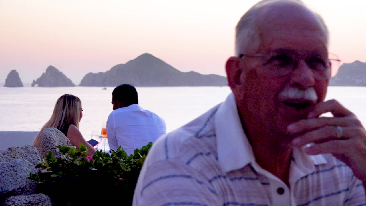 A senior man with glasses and a mustache sits close to camera at a beachside restaurant while diners face the water and mountains in the sunset background.