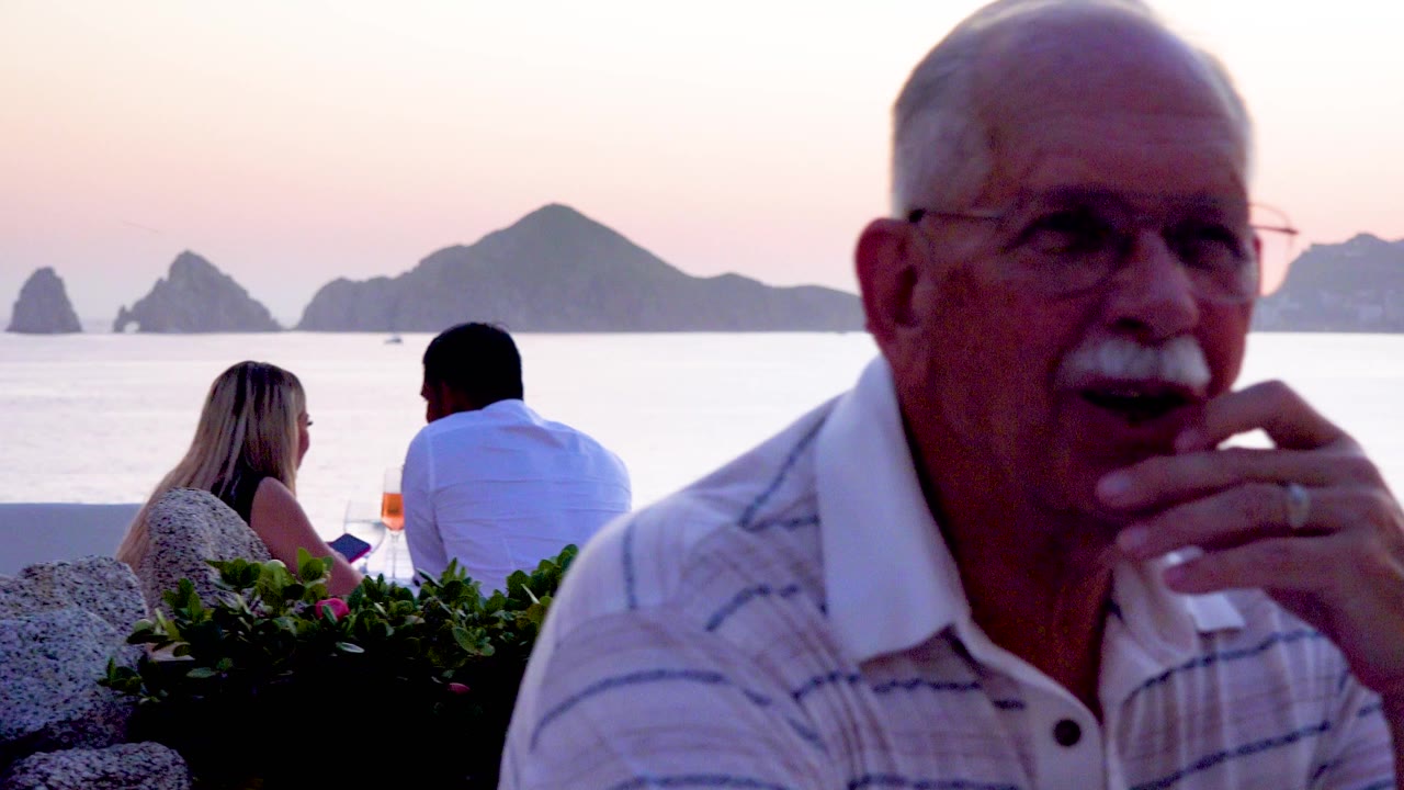 Senior man gestures near his mouth at a seaside restaurant table.