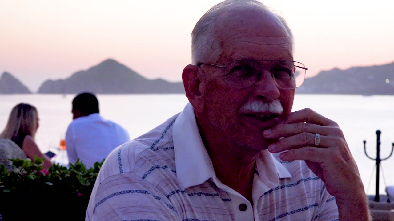 The man turns slightly to the side with his hand near his face, framed against silhouetted mountains, water, and a candlelit restaurant table.