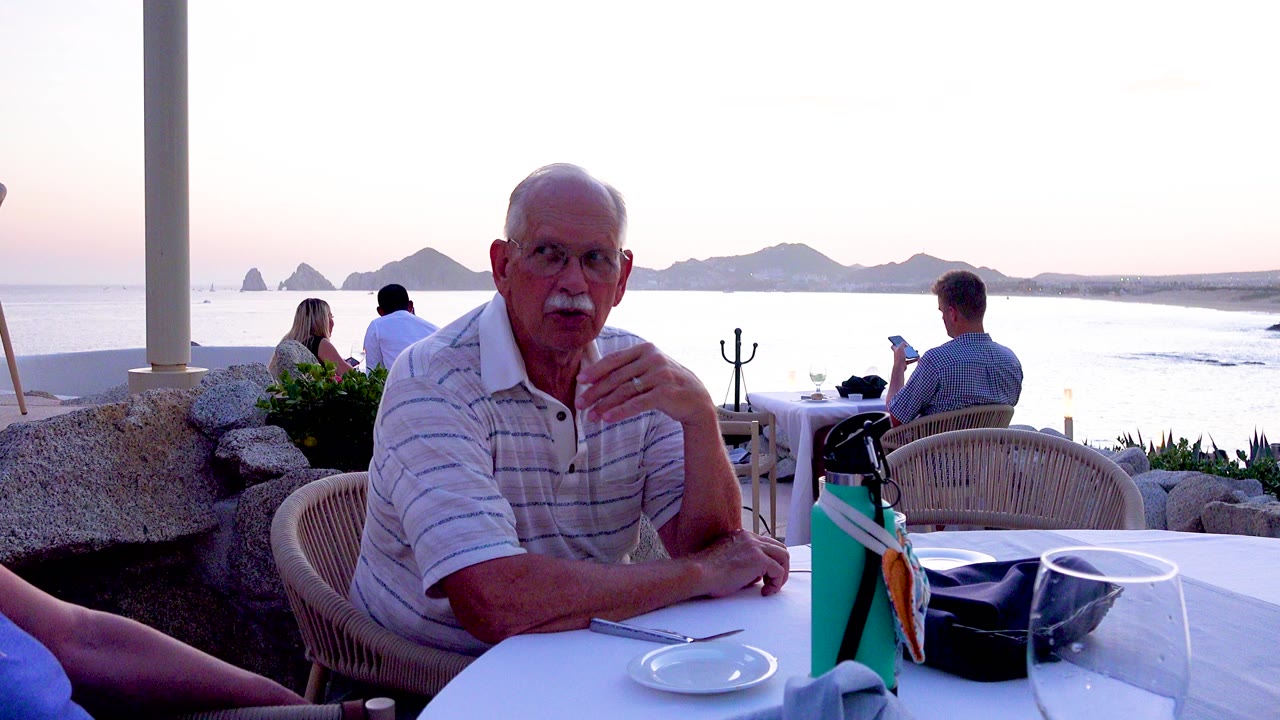 A senior man with glasses and a white mustache sits at a white patio table, with other diners and a bright coastal horizon behind him.
