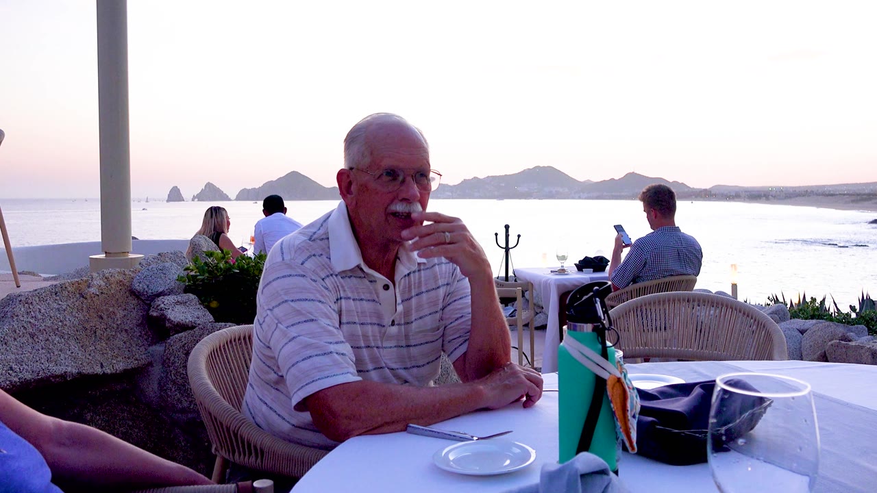 The seated man lifts his hand near his mouth, with the white tableware in front of him and silhouettes of islands across the water behind the patio.
